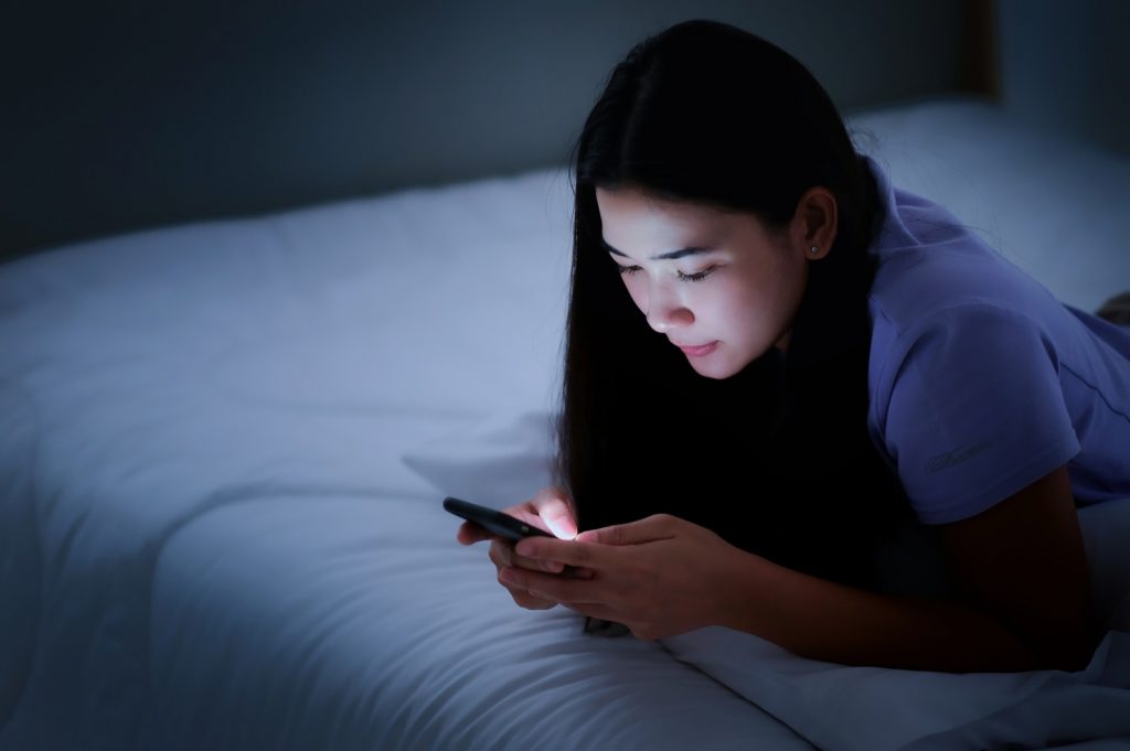 Female lying on the white bed and playing smartphones during night time.