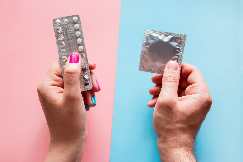 A woman holding the contraceptive pill and a man holding a condom on a pink and a blue background 