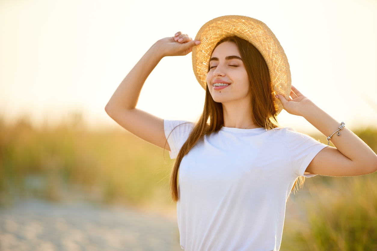 Spa wellness beach beauty woman relaxing and sun bathing on beach in straw hat. Beautiful serene and peaceful young female model with teeth braces on holiday travel resort enjoting vacation. Flower in braided hair.