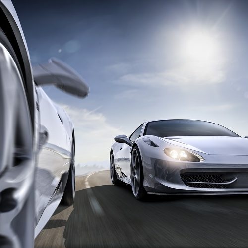 Low angle extreme close up view of two generic sports cars driving at speed along flat road in winter under bright blue sky with intentional lens flare.